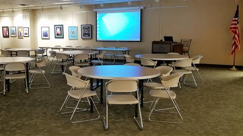 Waugaman Gallery complete with circular tables with seating at each table, projector and projection screen, lectern and rectangular table with seating
