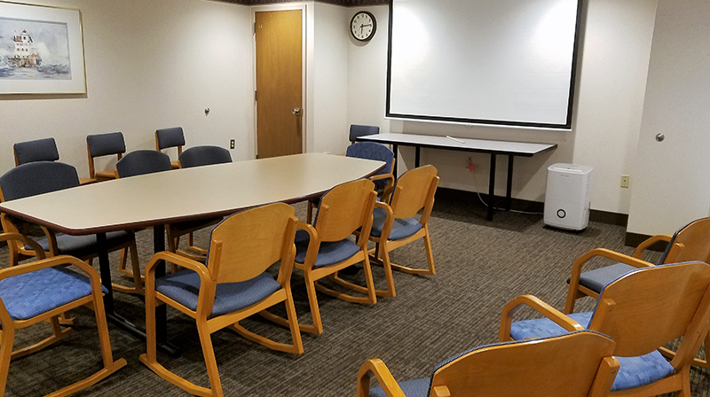 Room image showing conference table with seating on all sides, large projection screen, and additional chairs lining the walls
