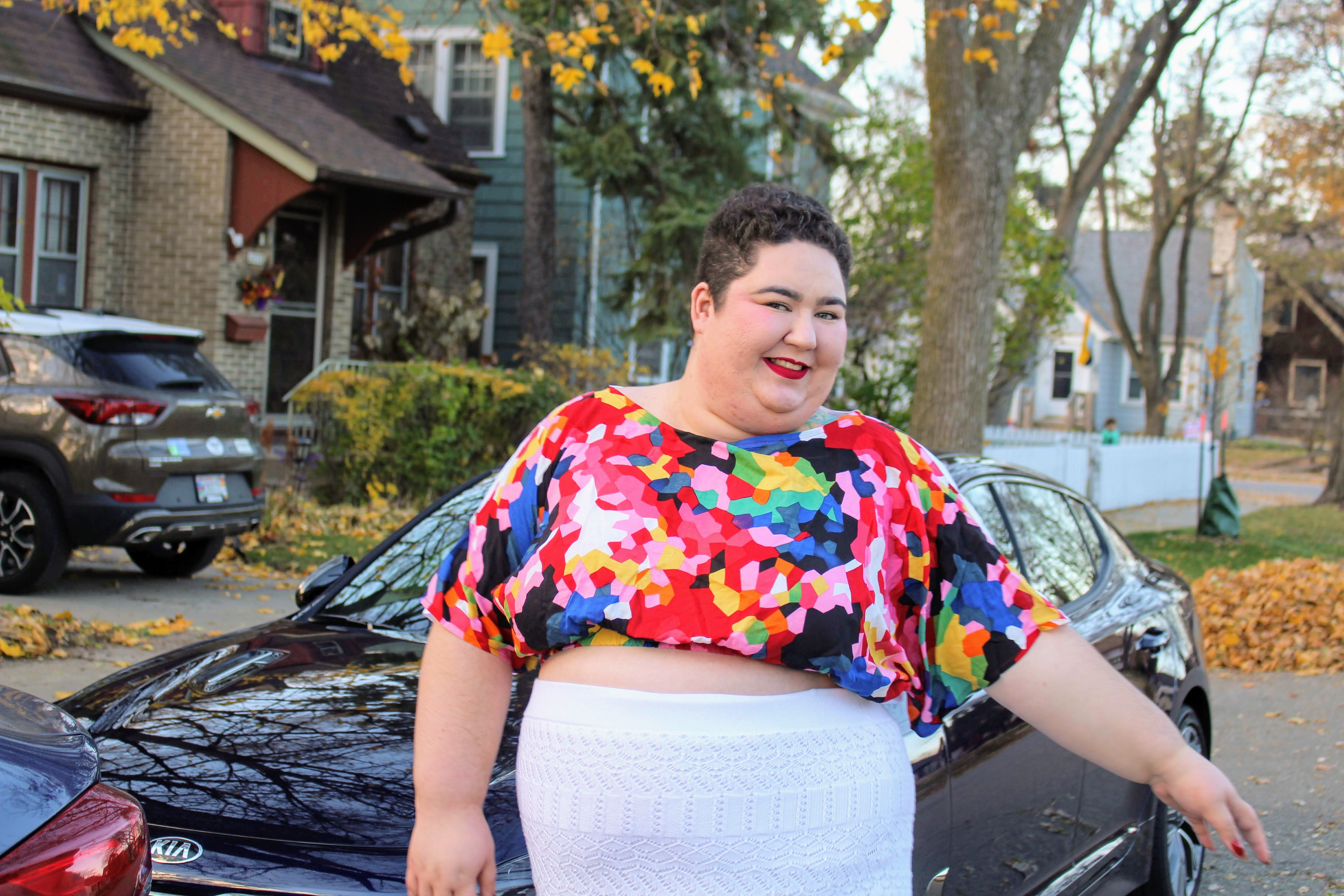A photo of Chaya Milchtein standing in front of a car outside. She's very stylish in a multicolor blouse and bright white skirt.